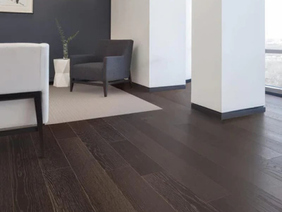 A modern living room features Commercial Hardwood planks in dark wood, a white rug, gray and white armchairs, and a white vase with green branches against a dark accent wall near large windows.