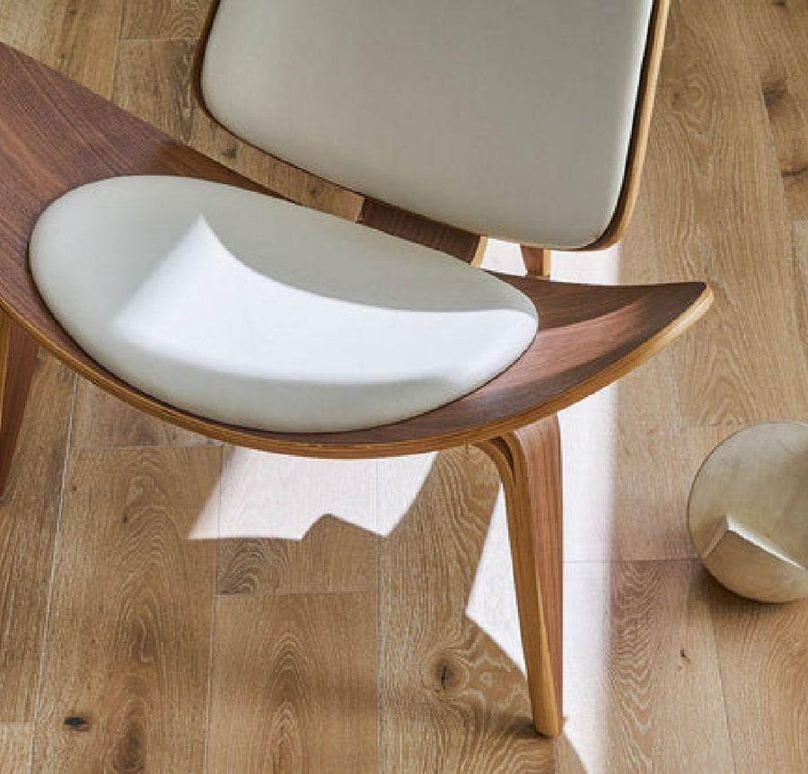 A modern wooden chair with a light beige cushioned seat and backrest is placed on Commercial Hardwood flooring next to a small metallic cylinder, while sunlight and shadows highlight the scene’s contrast.
