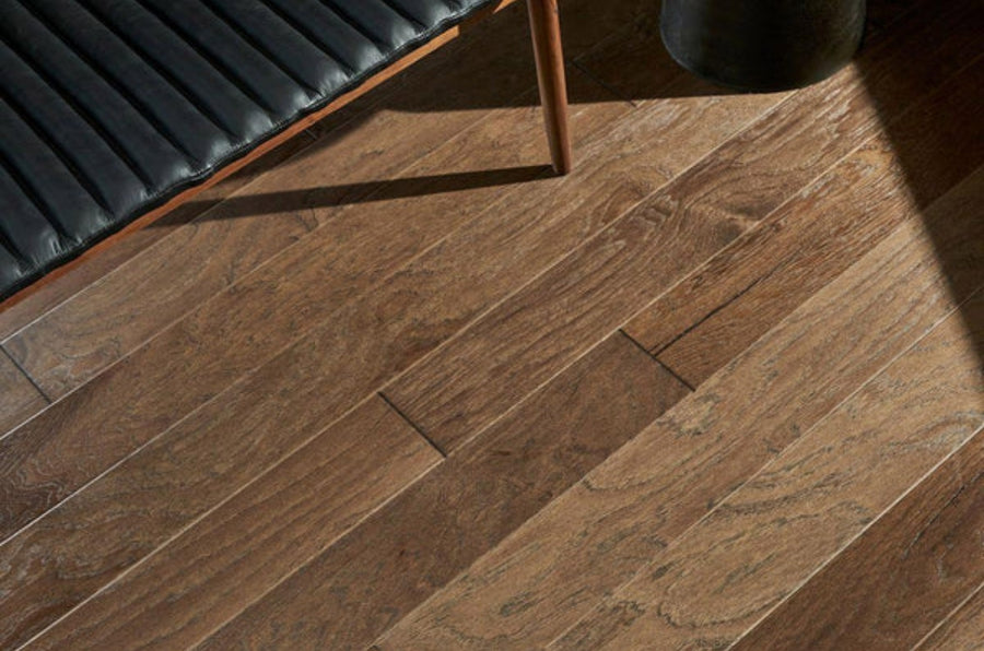 A close-up of Commercial Hardwood flooring in light brown with visible grain patterns, shown beside a black cushioned bench with wooden legs and a round black object.