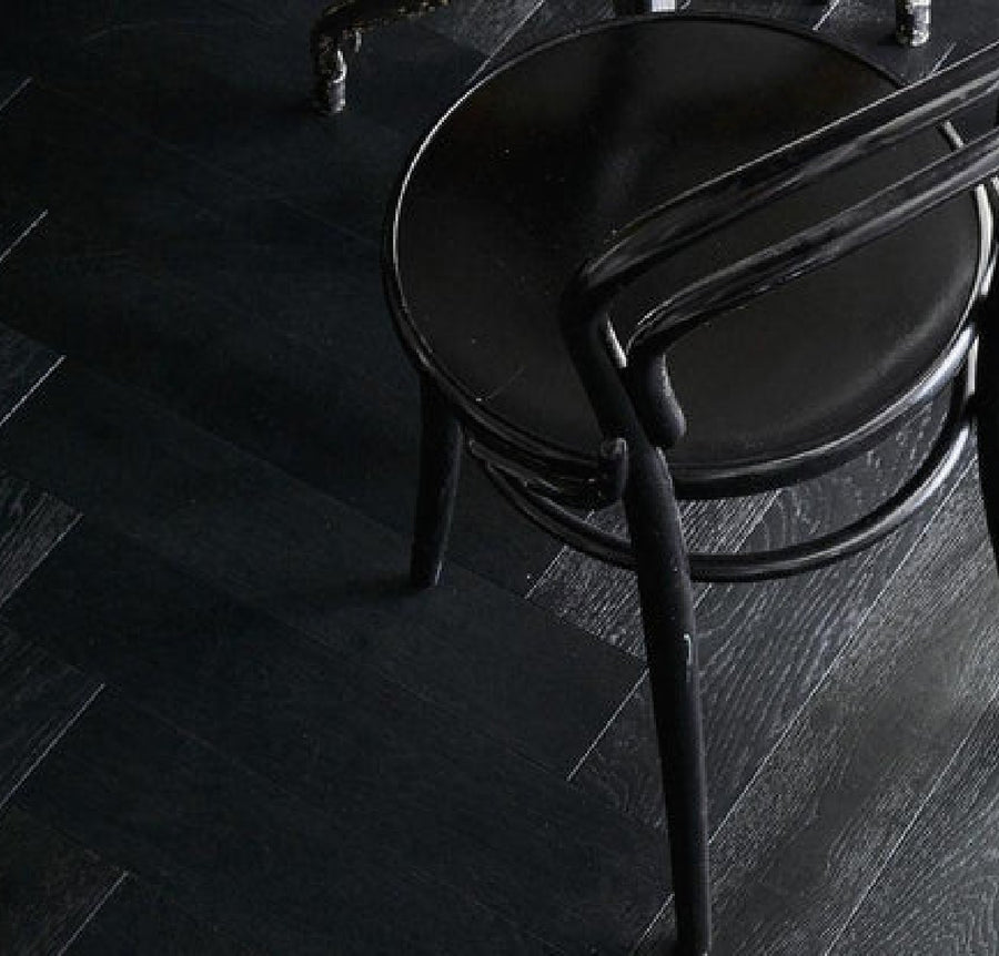A black round chair with a curved backrest sits on Commercial Hardwood flooring in a dark herringbone pattern. The minimal, monochromatic scene reveals only part of the chair and the floor.