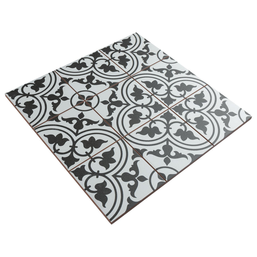 A square ceramic tile with a black and white vintage-style intricate floral and geometric pattern, divided into nine smaller squares with thin, brown grid lines.