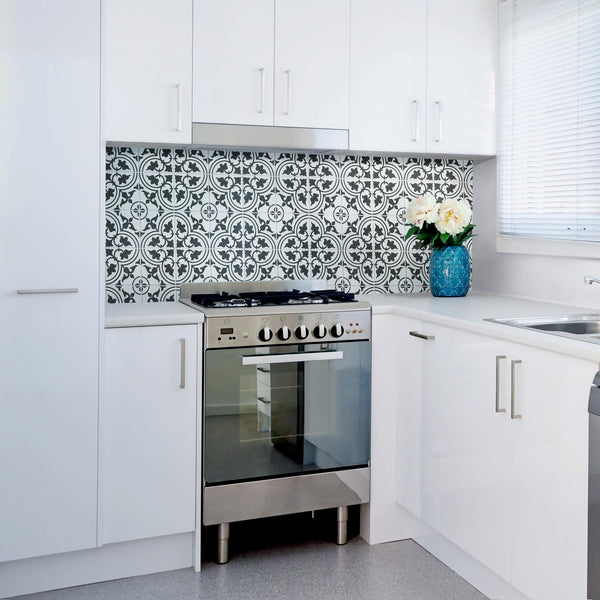 A modern white kitchen with a stainless steel oven, patterned black-and-white tile backsplash, white cabinets, and a blue vase with white flowers on the counter near a window with blinds.