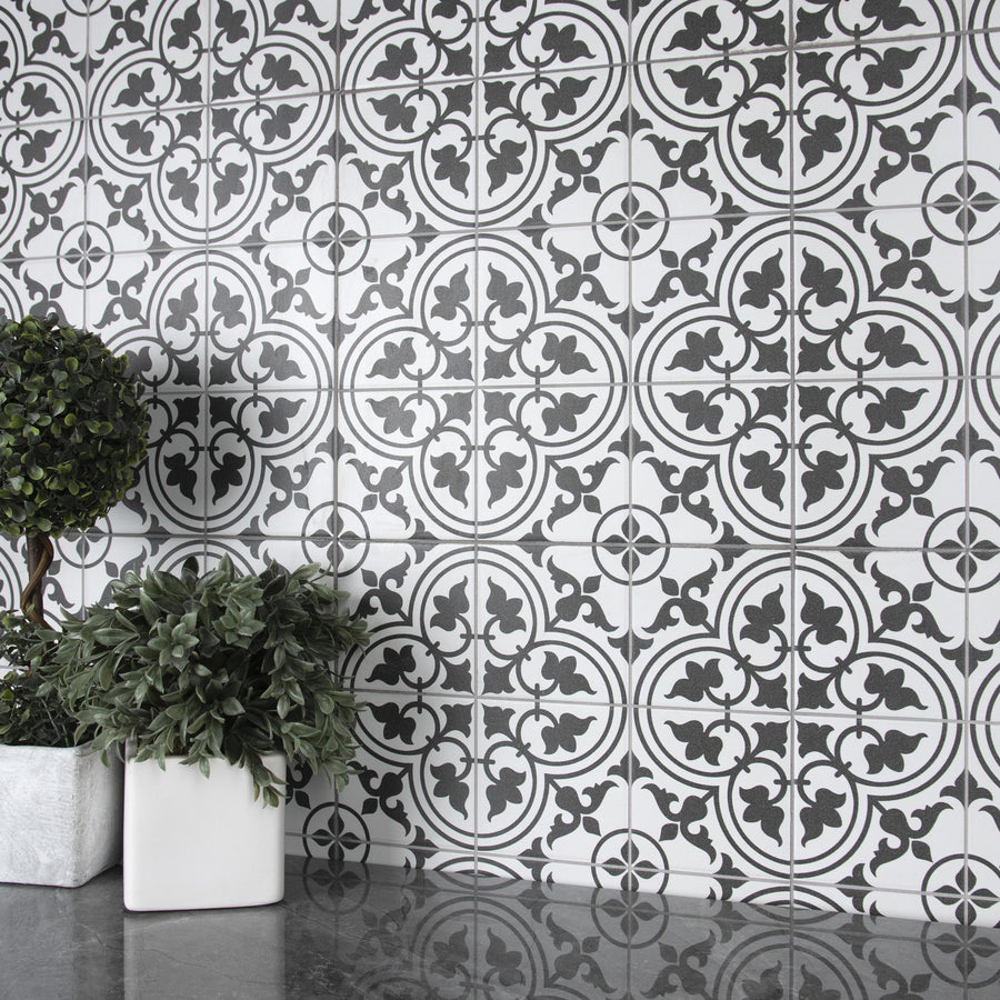 Black and white patterned tiles create an ornate backdrop. Two potted plants with green leaves, one in a silver square pot and one in a white square pot, sit on a reflective gray surface in front of the tiles.
