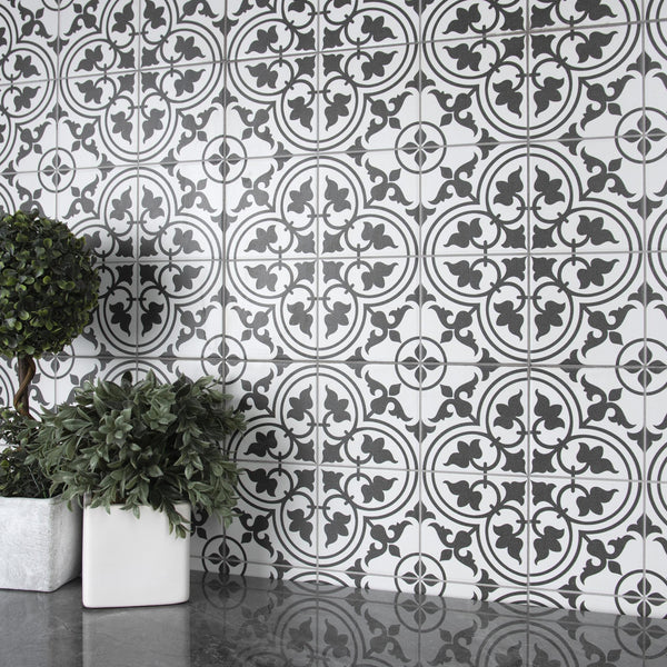 Black and white patterned tiles create an ornate backdrop. Two potted plants with green leaves, one in a silver square pot and one in a white square pot, sit on a reflective gray surface in front of the tiles.