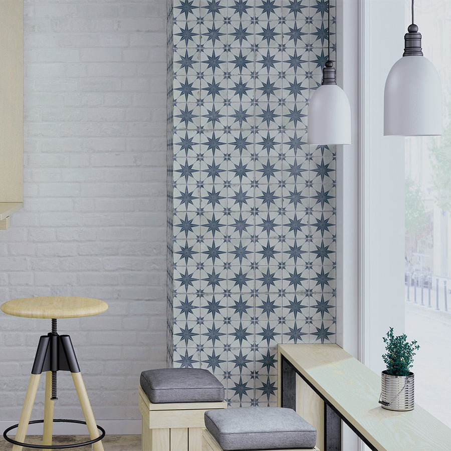 Modern interior with white brick walls, patterned blue-and-white accent wall, wooden stool, cushioned bench seating, small potted plant, and two hanging white pendant lights near large windows.