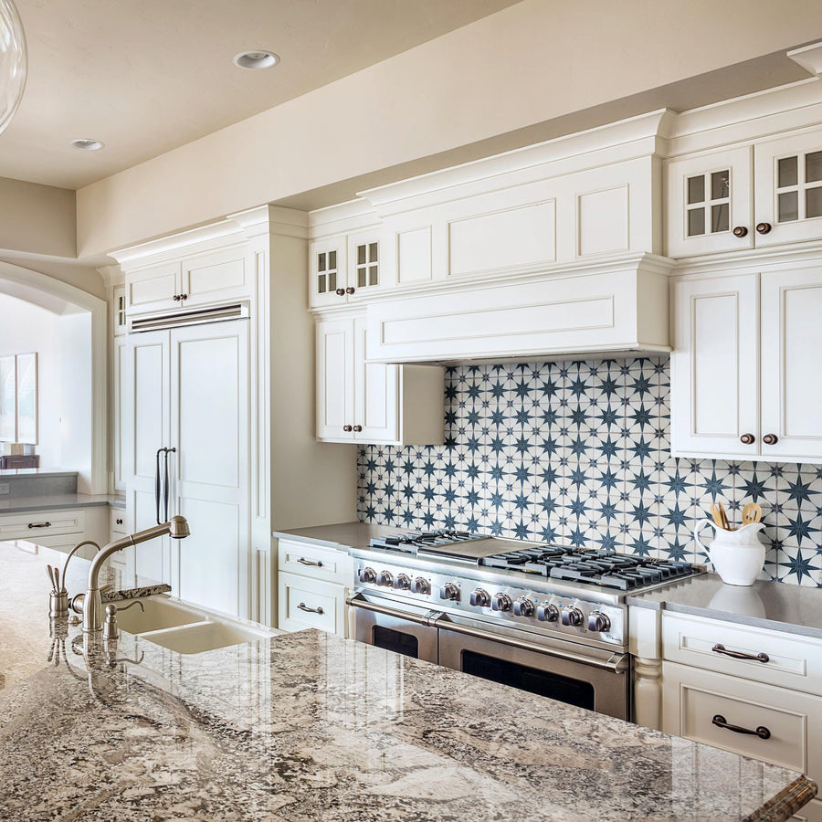 A bright, modern kitchen with white cabinets, a patterned blue and white tile backsplash, stainless steel stove, built-in fridge, marble countertops, and a sink with a gooseneck faucet.