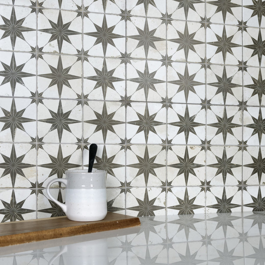 A white and gray patterned tile backsplash with star designs, a white ceramic mug with a black spoon, and a wooden board on a glossy white countertop.