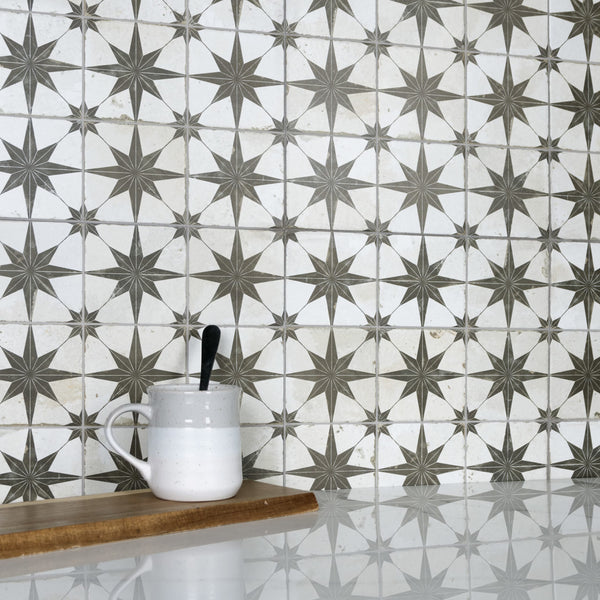 A white and gray patterned tile backsplash with star designs, a white ceramic mug with a black spoon, and a wooden board on a glossy white countertop.