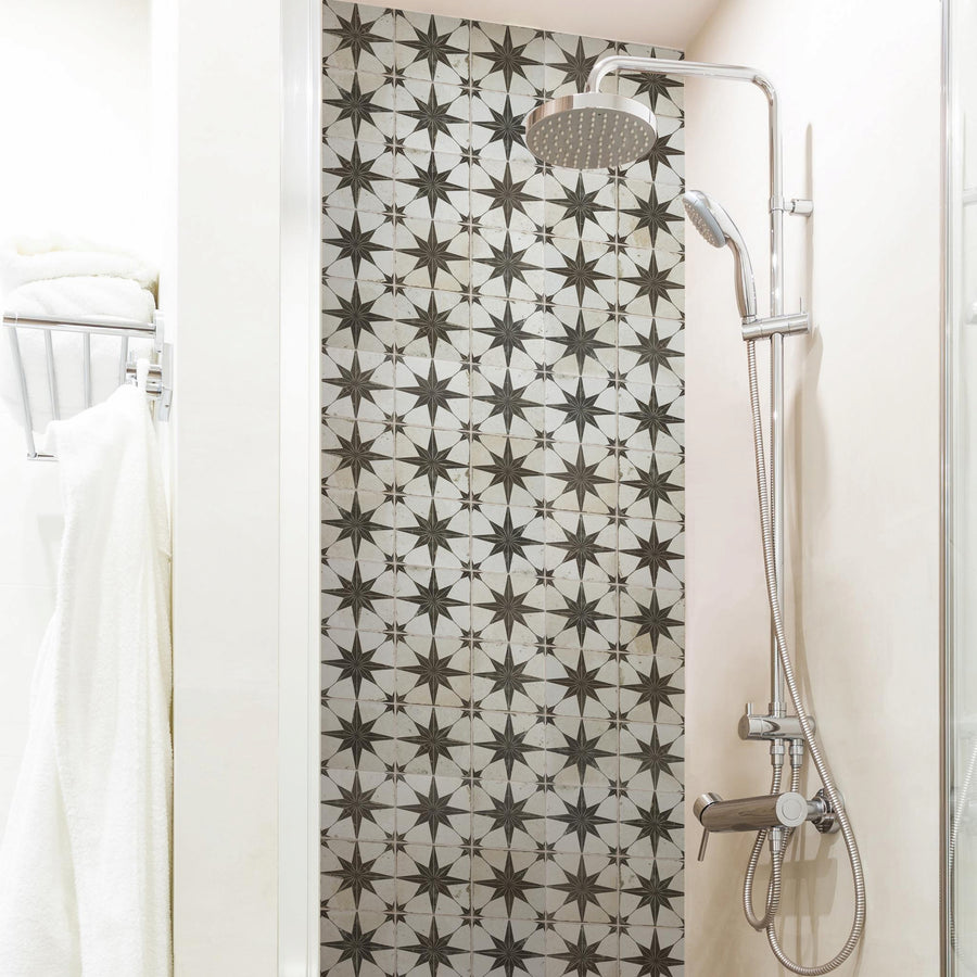 A modern shower with a rain showerhead and a handheld shower, featuring a wall of black and white geometric star-patterned tiles. White towels hang on a rack to the left.