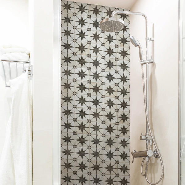 A modern shower with a rain showerhead and a handheld shower, featuring a wall of black and white geometric star-patterned tiles. White towels hang on a rack to the left.