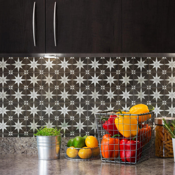 A kitchen counter with a metal pot of green herbs, a wire basket holding colorful bell peppers, and a few limes and lemons in front. The backsplash has a geometric star pattern, and dark cabinets hang above.