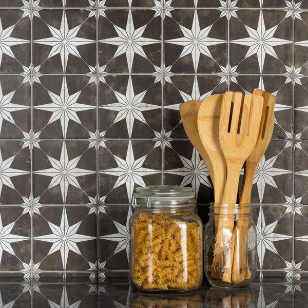 A glass jar of fusilli pasta and a jar holding wooden cooking utensils sit on a black countertop in front of a black and white star-patterned tile backsplash.