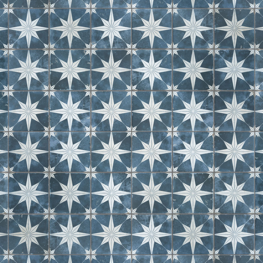 A patterned tile surface featuring repeating white eight-pointed stars on a blue background, with each star centered in a square, creating a geometric, symmetrical design.