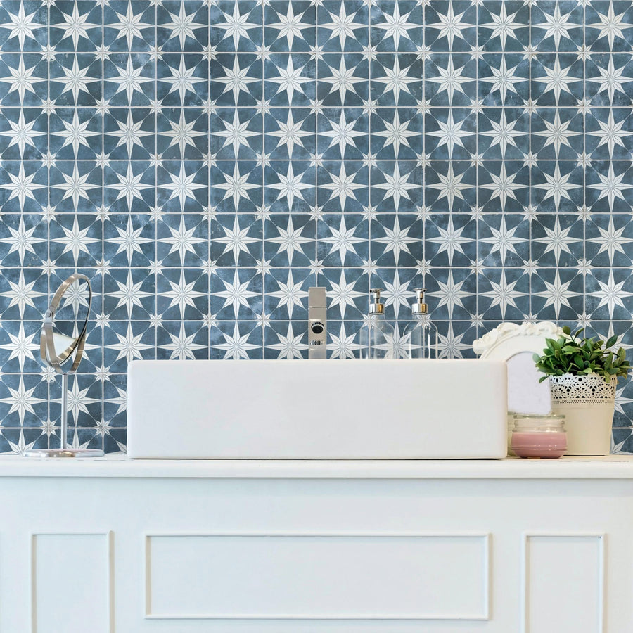 A white bathroom sink with a modern faucet sits on a white counter. Behind it, the wall is covered with blue and white star-patterned tiles. There are soap dispensers, a potted plant, and a round mirror on the counter.