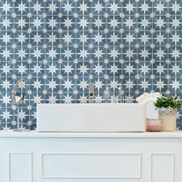 A white bathroom sink with a modern faucet sits on a white counter. Behind it, the wall is covered with blue and white star-patterned tiles. There are soap dispensers, a potted plant, and a round mirror on the counter.