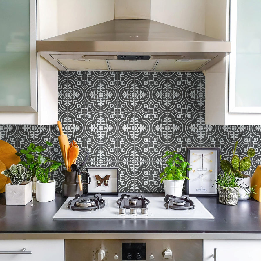 A modern kitchen with a black and white patterned tile backsplash, gas stove, stainless steel range hood, and countertop decorated with potted plants, framed art, knives, and utensils.