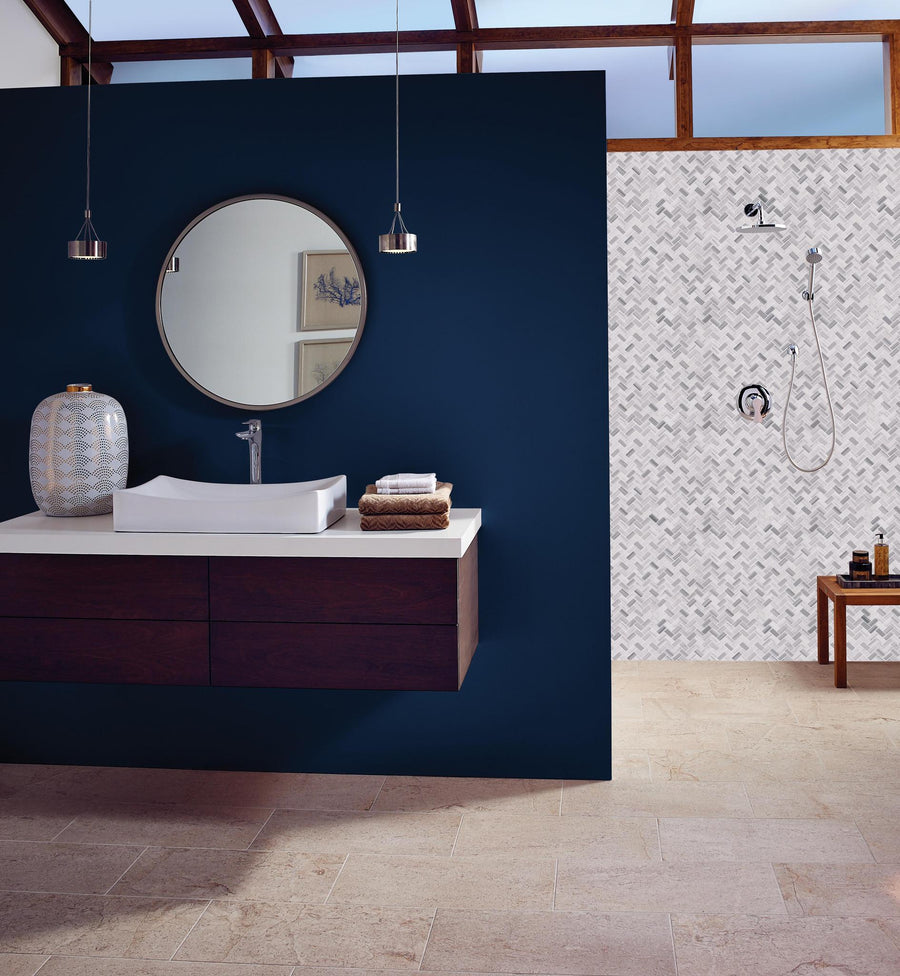 Modern bathroom with a dark blue accent wall, round mirror, floating vanity with a vessel sink, and pendant lights. The shower area features white herringbone tiles and a wooden stool with bath items.