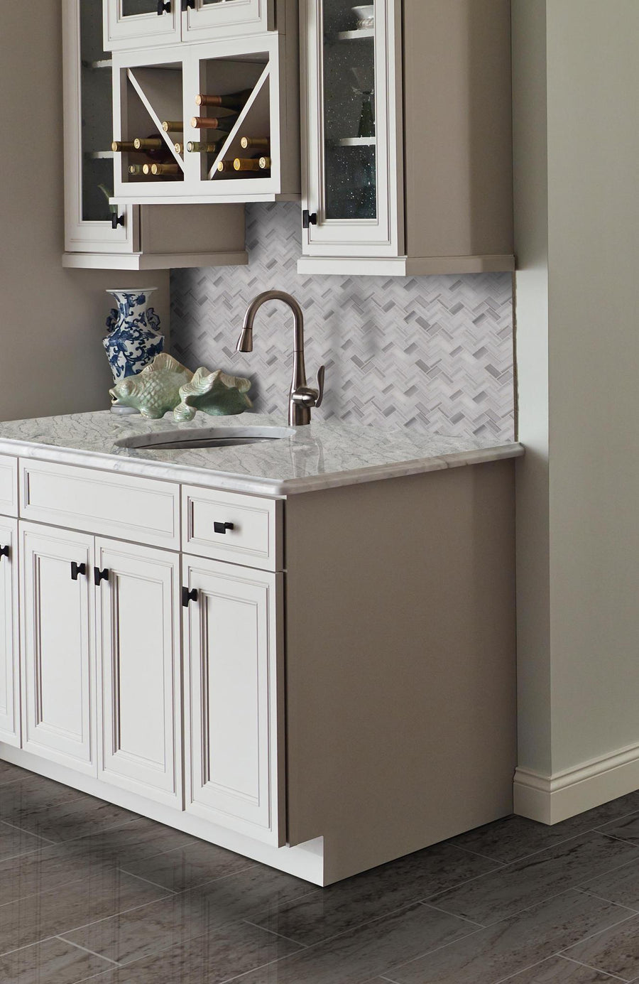 A kitchen corner with light cabinets, a marble countertop, a silver faucet, a blue and white vase, decorative shells, and a gray and white herringbone tile backsplash.