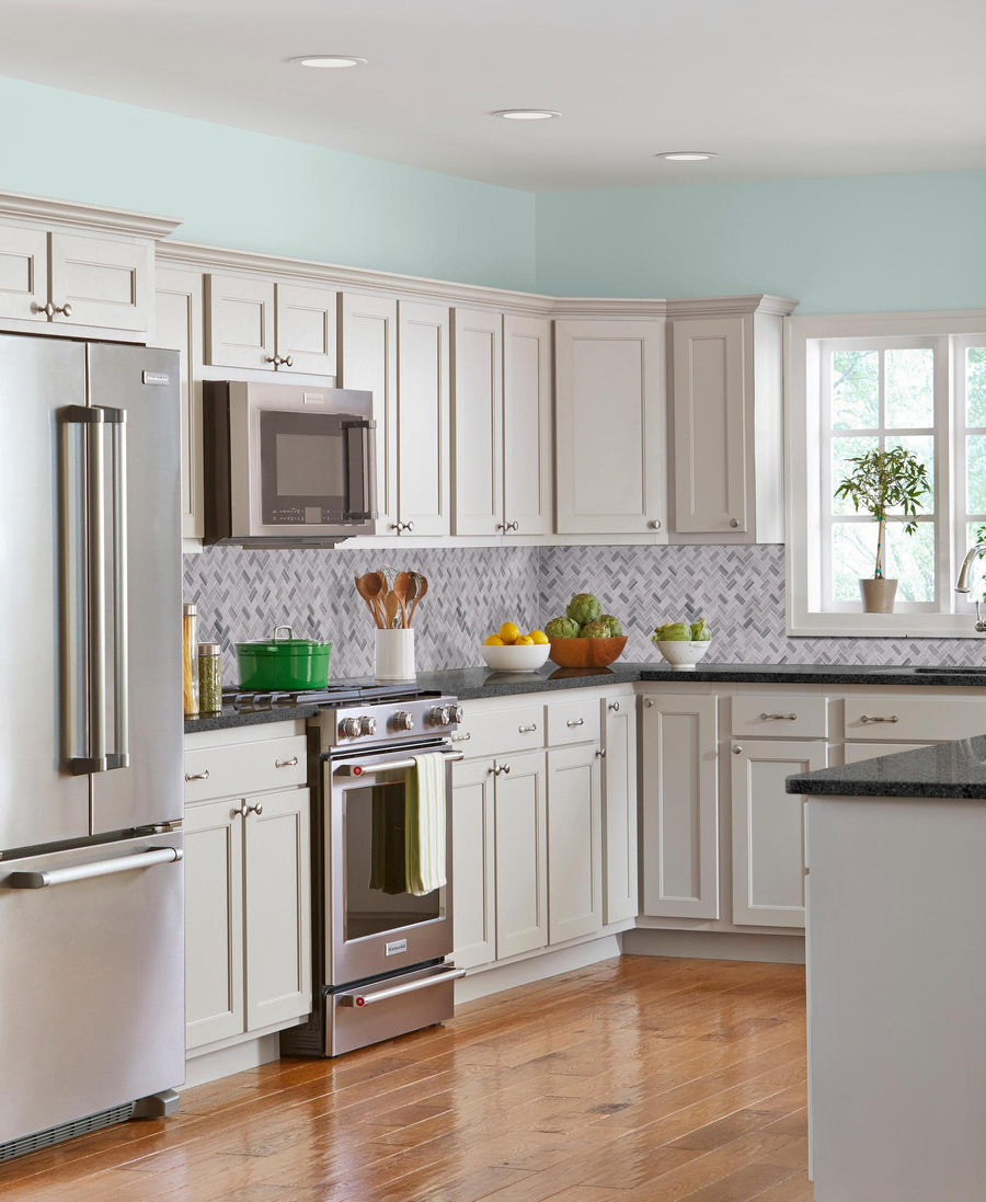 Modern kitchen with white cabinets, stainless steel appliances, wooden floor, and a gray patterned backsplash. The counters display a plant, utensils, produce bowls, and a green pot on the stove near a window.