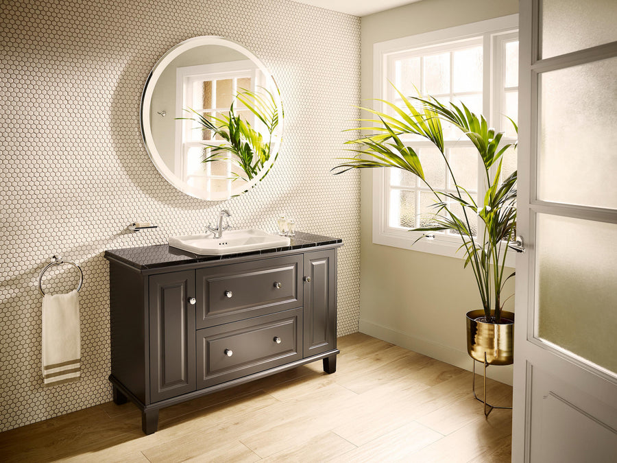 Modern bathroom with a black vanity, round mirror, potted palm plant, hexagonal tile wall, wooden floor, and natural light streaming through a window. A hand towel hangs on a ring beside the vanity.
