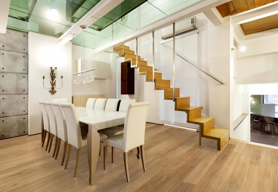 Modern dining room with a long white table, eight upholstered chairs, wooden floors, and a floating wooden staircase with a metal railing leading to a glass mezzanine. The space is bright with minimalist decor.