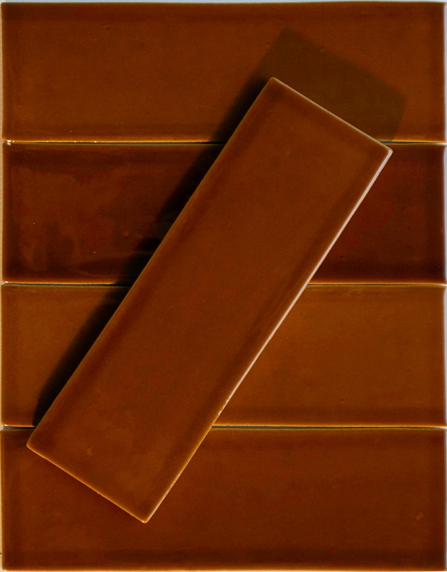 A Noir Crackle 2.5x8 Ceramic Tile in the shade "Honey I’m Home" rests diagonally across four aligned tiles, showcasing their glossy surfaces and warm hues against a rich brown backdrop for a decadent, layered look.