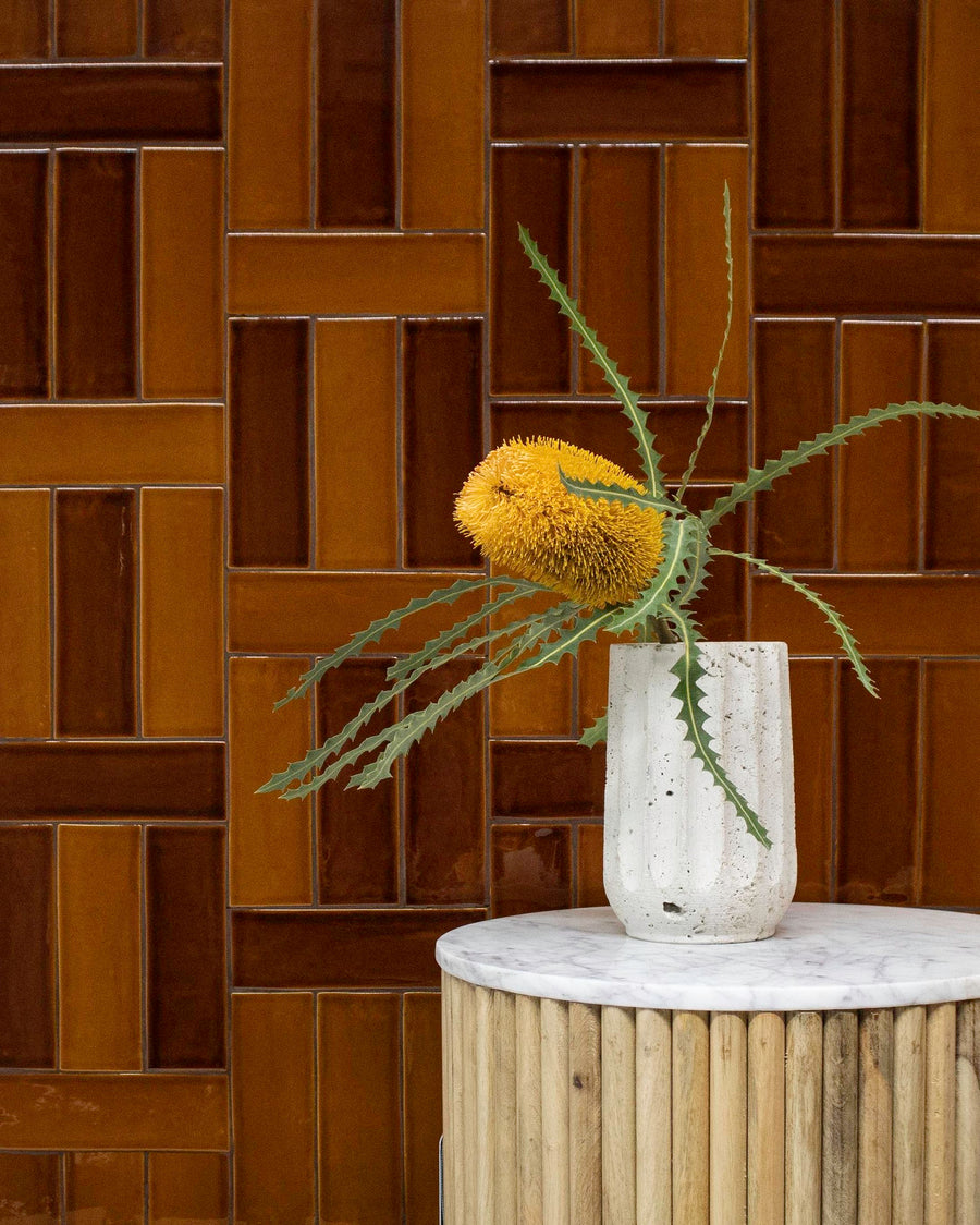A single yellow protea flower with jagged green leaves in a white vase sits on a small, round, marble-topped table with a wood base. The background features glossy brown tiles in a geometric pattern.