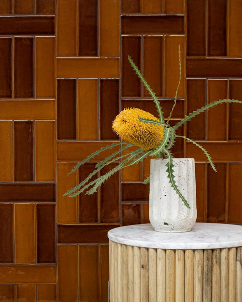 A single yellow protea flower with jagged green leaves in a white vase sits on a small, round, marble-topped table with a wood base. The background features glossy brown tiles in a geometric pattern.