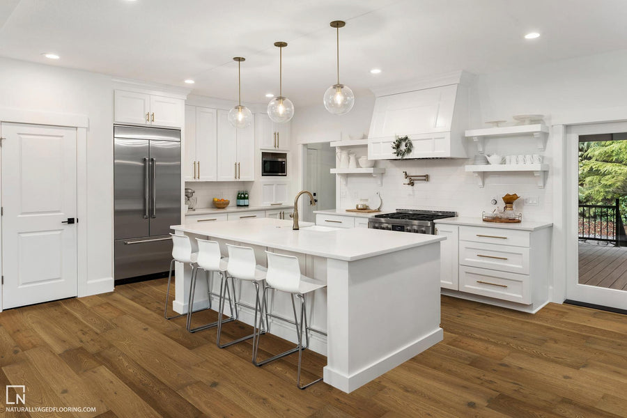 A modern white kitchen features an island with four white barstools, stainless steel appliances, pendant lights, open shelves with dishes, and large windows overlooking greenery. The floor is light wood.