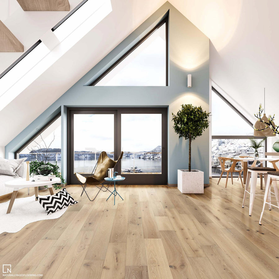 A modern, bright attic living space with large windows, light wood flooring, a potted tree, and contemporary furniture. The windows reveal a scenic view of water, hills, and buildings outside.