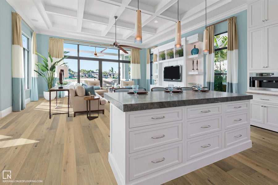 A bright, modern kitchen with a large island, white cabinets, pendant lights, wood flooring, and an open living area with large windows overlooking palm trees and water.