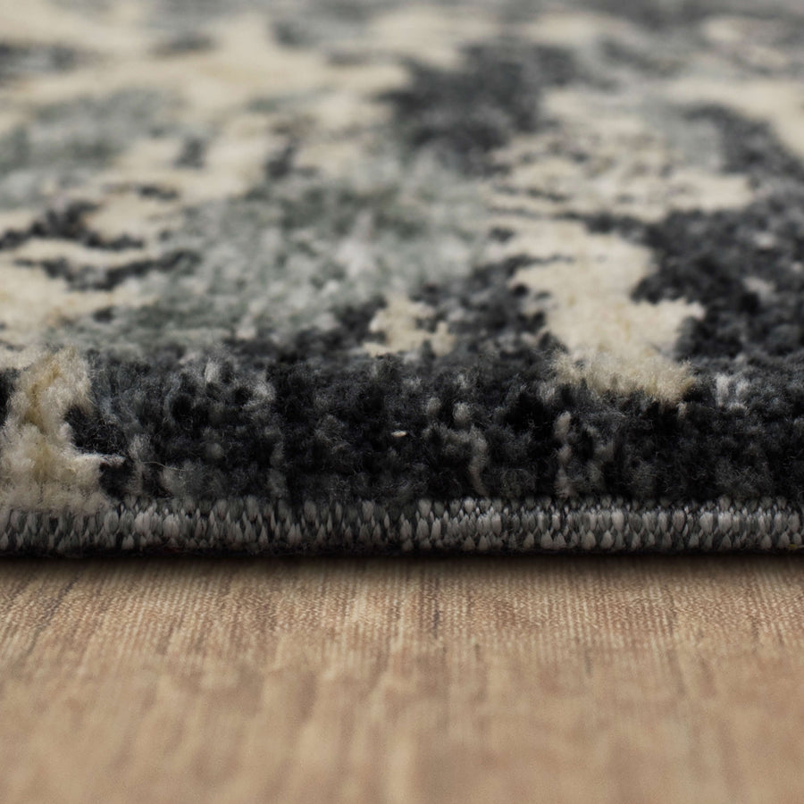 Close-up side view of a rug with a thick, textured black, white, and gray pattern, lying flat on a light wooden floor.