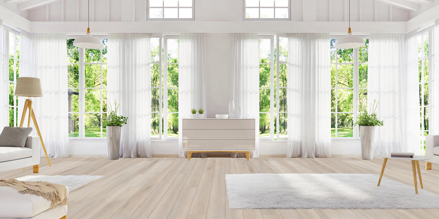 Bright, spacious living room with large windows, sheer white curtains, light wood floors, white walls, minimal furniture, and green trees visible outside. Decor includes rugs, lamps, plants, and a dresser with vases.