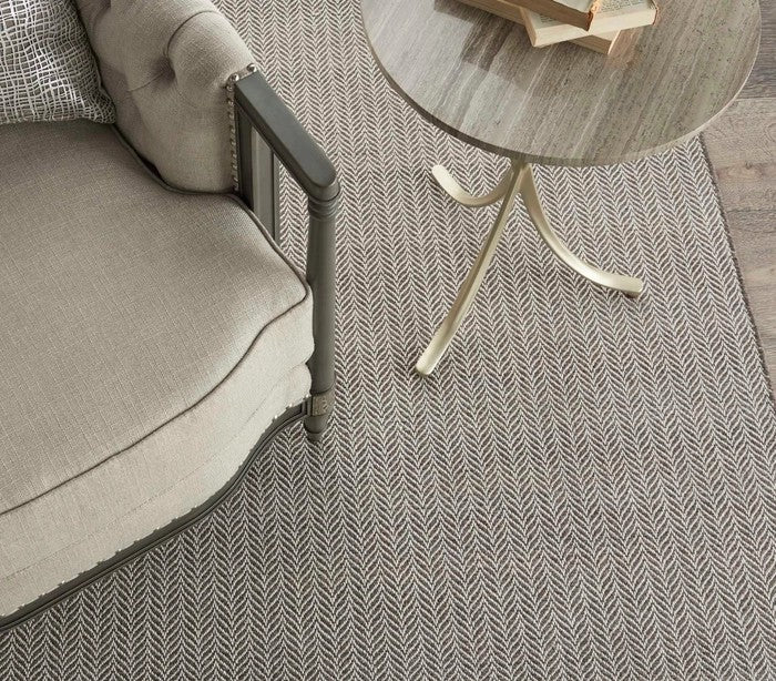 A beige upholstered armchair sits next to a round, marble-topped side table with gold legs on a textured, gray herringbone-patterned rug. A book and glasses rest on the table.
