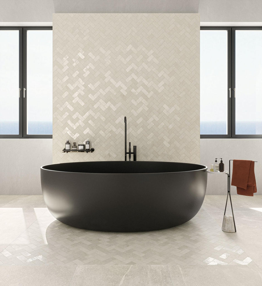 A modern bathroom with a large, round black bathtub, light herringbone tile wall, minimalist fixtures, two windows, and a brown towel on a hook. Natural light fills the space.