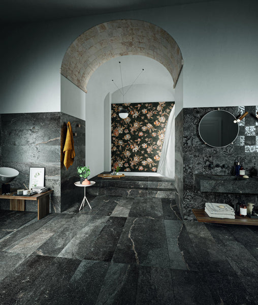 A modern bathroom with dark stone tiles, a raised bathtub with floral wallpaper behind it, a round mirror above a wooden shelf with toiletries, and a yellow towel hanging on the wall.