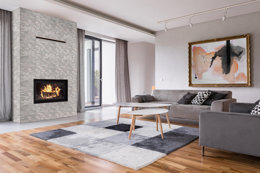 Modern living room with gray sofas, a geometric rug, two coffee tables, a framed abstract painting, and a fireplace with a tiled surround. Large windows with curtains allow natural light into the room.