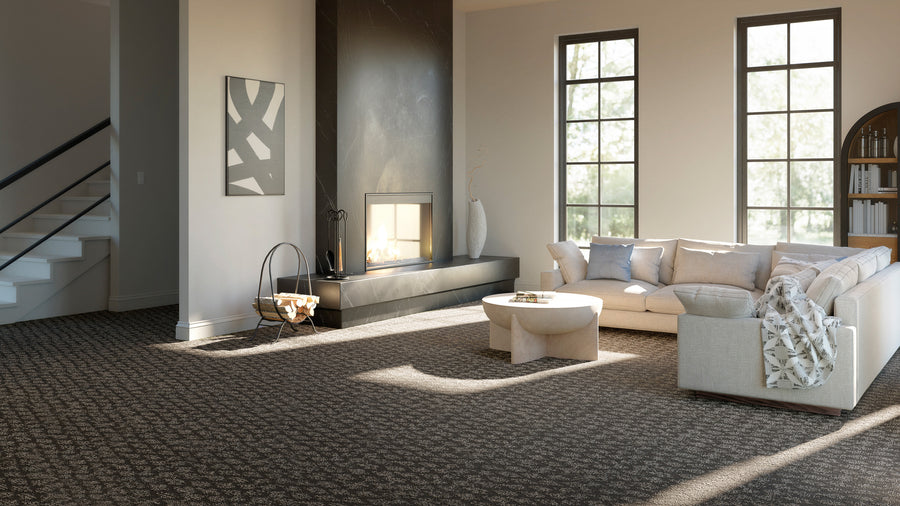 Modern living room with a large beige sectional sofa, round coffee table, fireplace, abstract wall art, and tall windows letting in natural light; dark patterned carpet covers the floor.