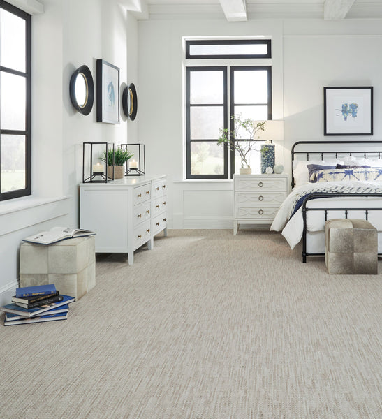 Bright bedroom with beige carpet, white walls, a black metal bed with white bedding, two white dressers, wall art, plants, and large windows letting in natural light. Two cube ottomans and books are on the floor.