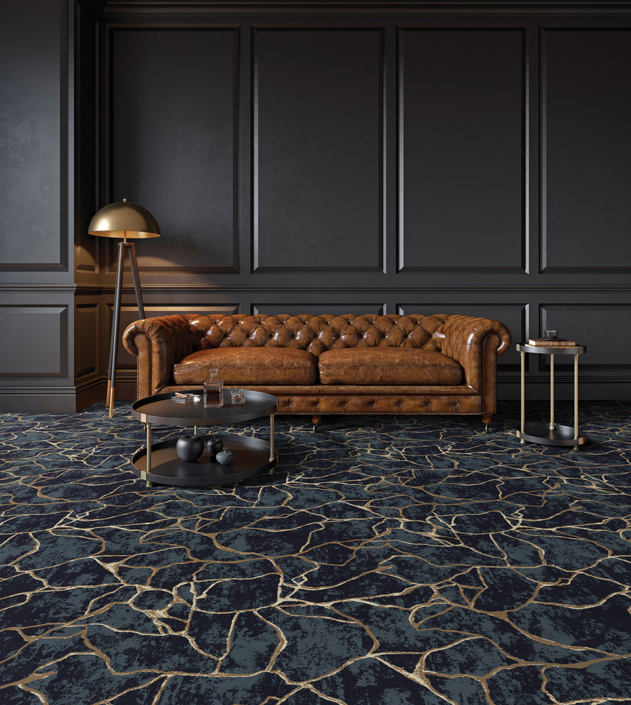 A stylish living room with dark paneled walls, a brown tufted leather sofa, a round glass coffee table, a small side table with books, a gold floor lamp, and a carpet with an abstract gold and dark pattern.