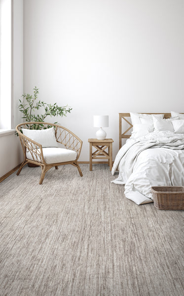 A bright, minimalist bedroom with a wicker chair, wooden bed, nightstand, white bedding, a small lamp, green plant, and a wicker basket on the light beige carpet. Sunlight streams in through a large window.
