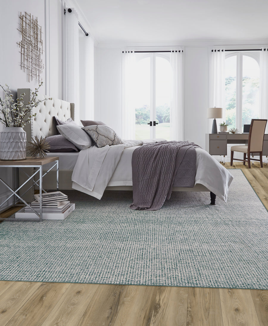Elegant bedroom with a tufted bed, gray bedding, and a lilac throw. Large windows let in natural light. A desk with a chair and lamps flank the room, and a textured rug covers part of the wood floor.