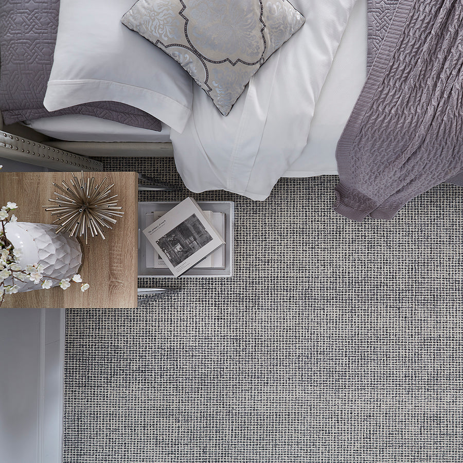 A top view of a neatly made bed with white and light grey bedding, a decorative pillow, a textured throw, a bedside table with a lamp, décor, and books, all on a patterned carpet.