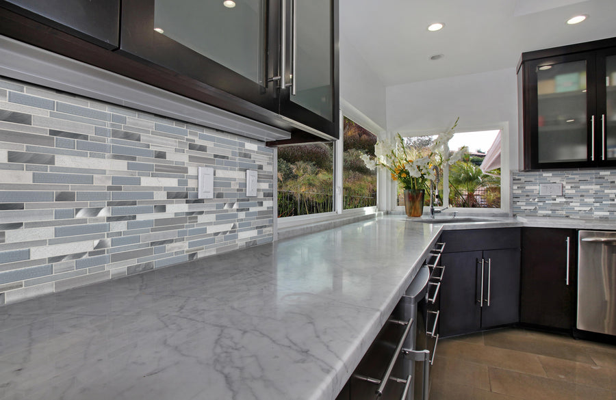 Modern kitchen with a long marble countertop, dark cabinets, stainless steel handles, and a gray mosaic tile backsplash. Large windows let in natural light; a vase with flowers sits on the counter near the windows.