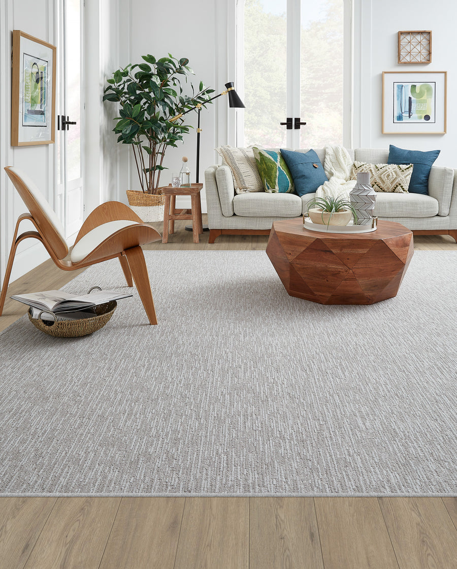 Modern living room with light wood floors, a textured gray rug, a geometric wooden coffee table, a cream sofa with blue and green pillows, wall art, a wooden accent chair, a plant, and large windows letting in natural light.