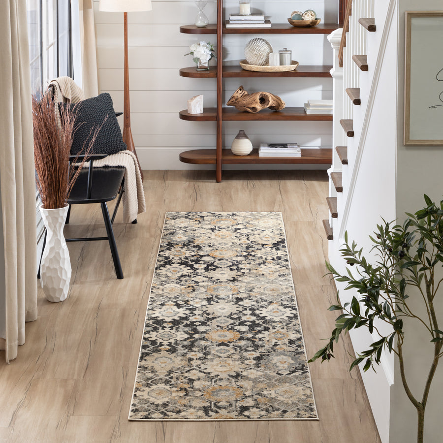 A hallway with light wood floors, a patterned runner rug, a black chair with a knit throw, a floor lamp, a potted plant, and wooden shelves displaying decor beside a staircase. Natural light fills the space.