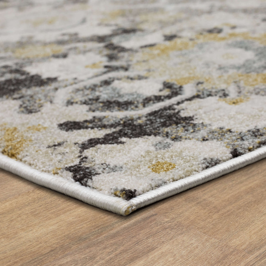 Close-up of a modern area rug with an abstract pattern in shades of white, gray, yellow, and black, placed on a light wood floor. The rug’s edge is neatly finished with a white border.