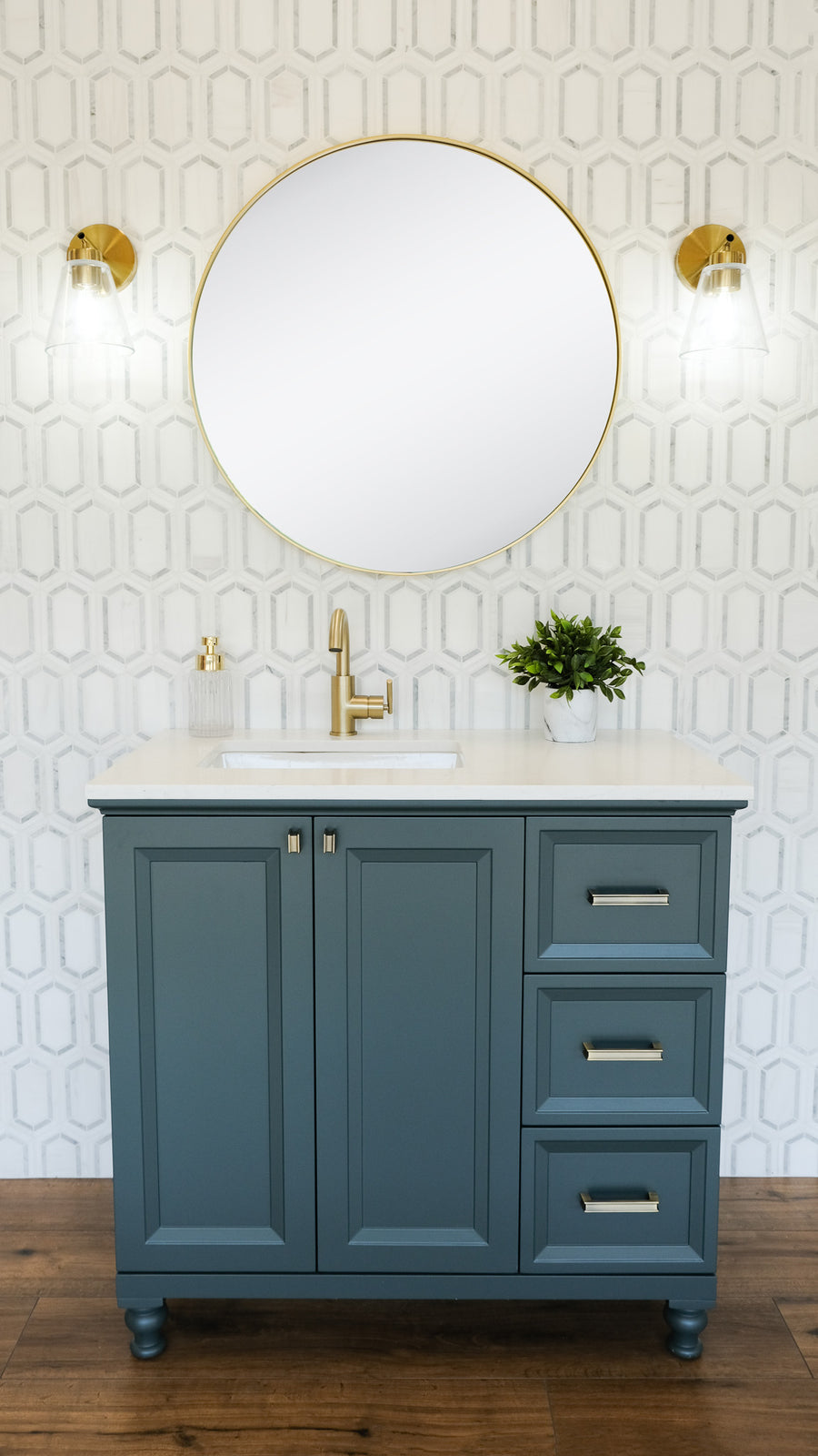 A modern bathroom vanity with a blue cabinet, brass hardware, white countertop, and built-in sink. Above is a large round mirror flanked by two brass wall sconces. A small plant and soap dispenser sit on the counter.