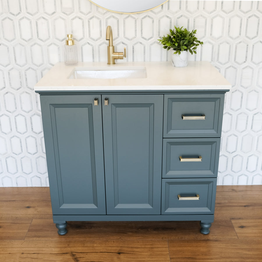 The Isabel 36" Vanity is a traditional bathroom piece with solid wood drawers, gold hardware, a white countertop, and an inset sink. Styled with geometric white tiles, a wood floor, soap dispenser, and potted plant for a finished look.