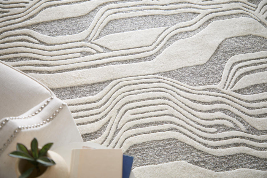 A close-up of a modern rug with raised, wavy cream lines on a textured gray background. Part of a light-colored armchair with nailhead trim and a small plant are visible in the corner.